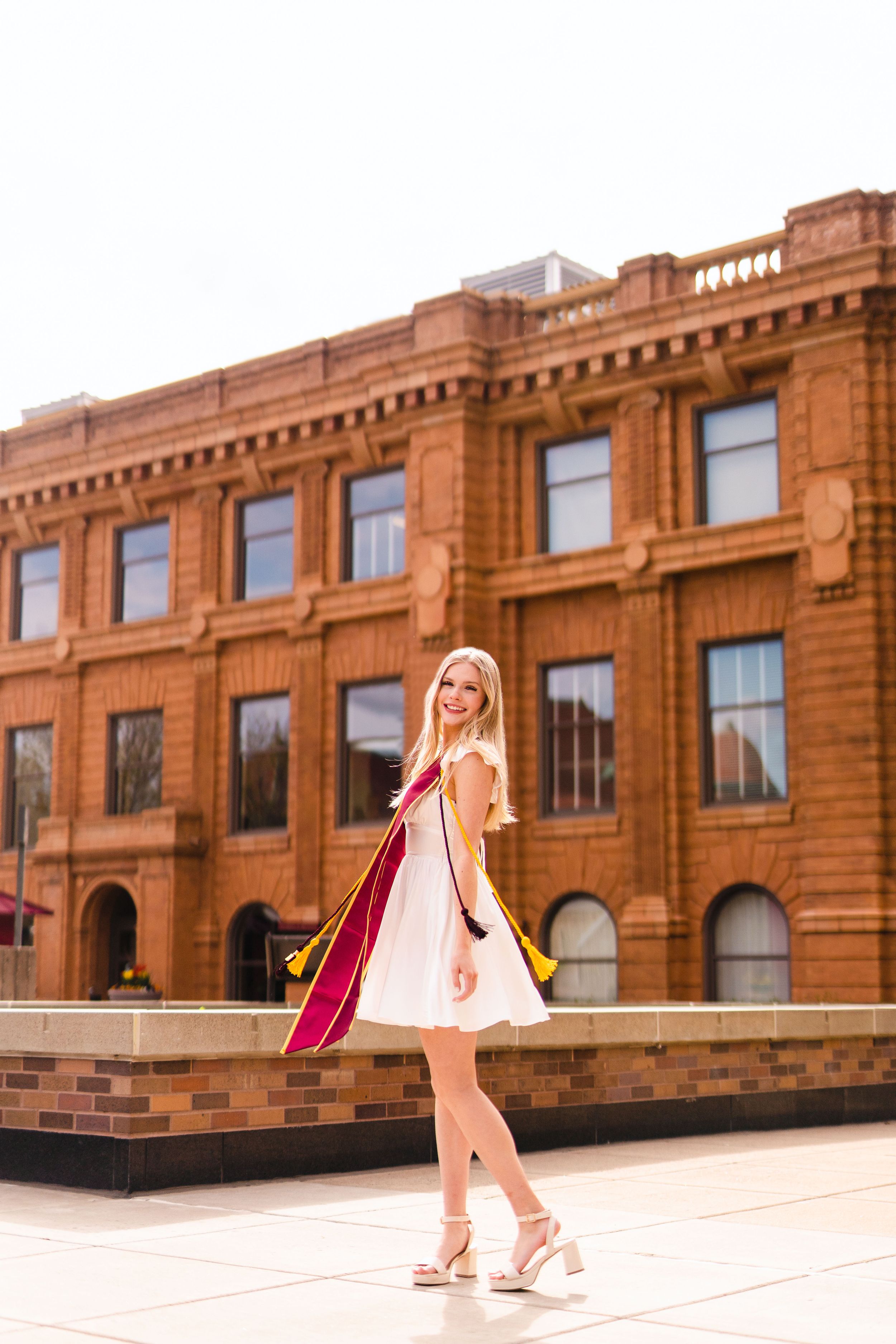 Graduate in white dress twirls on campus plaza near brick building.