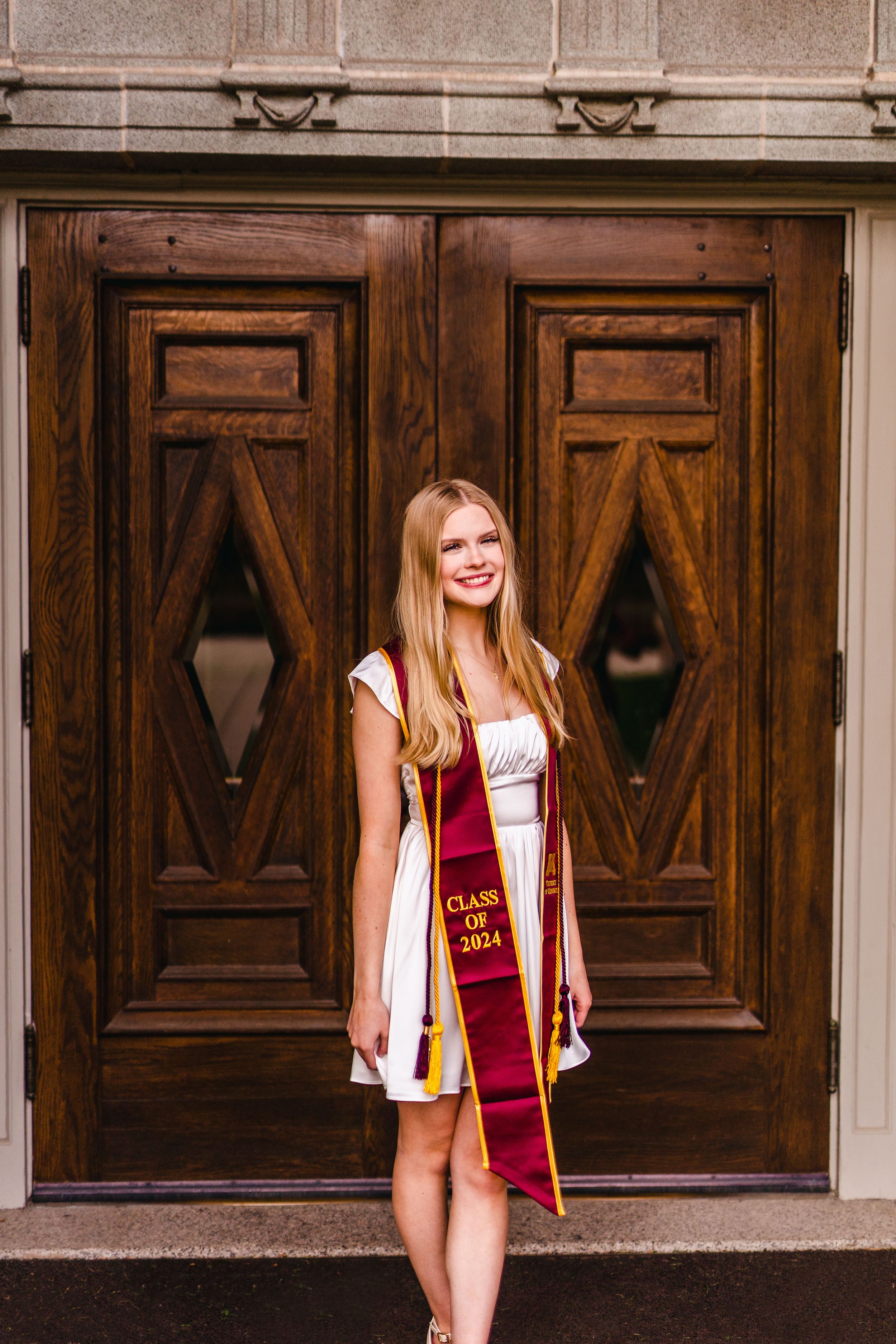 Graduate in a white dress and maroon graduation sash poses in front of ornate wooden doors.