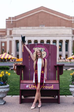 Graduate celebrates in a maroon UMN chair with cap raised in front of campus building.