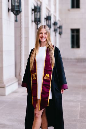 Graduate in academic regalia and white dress poses against stone wall with lanterns.