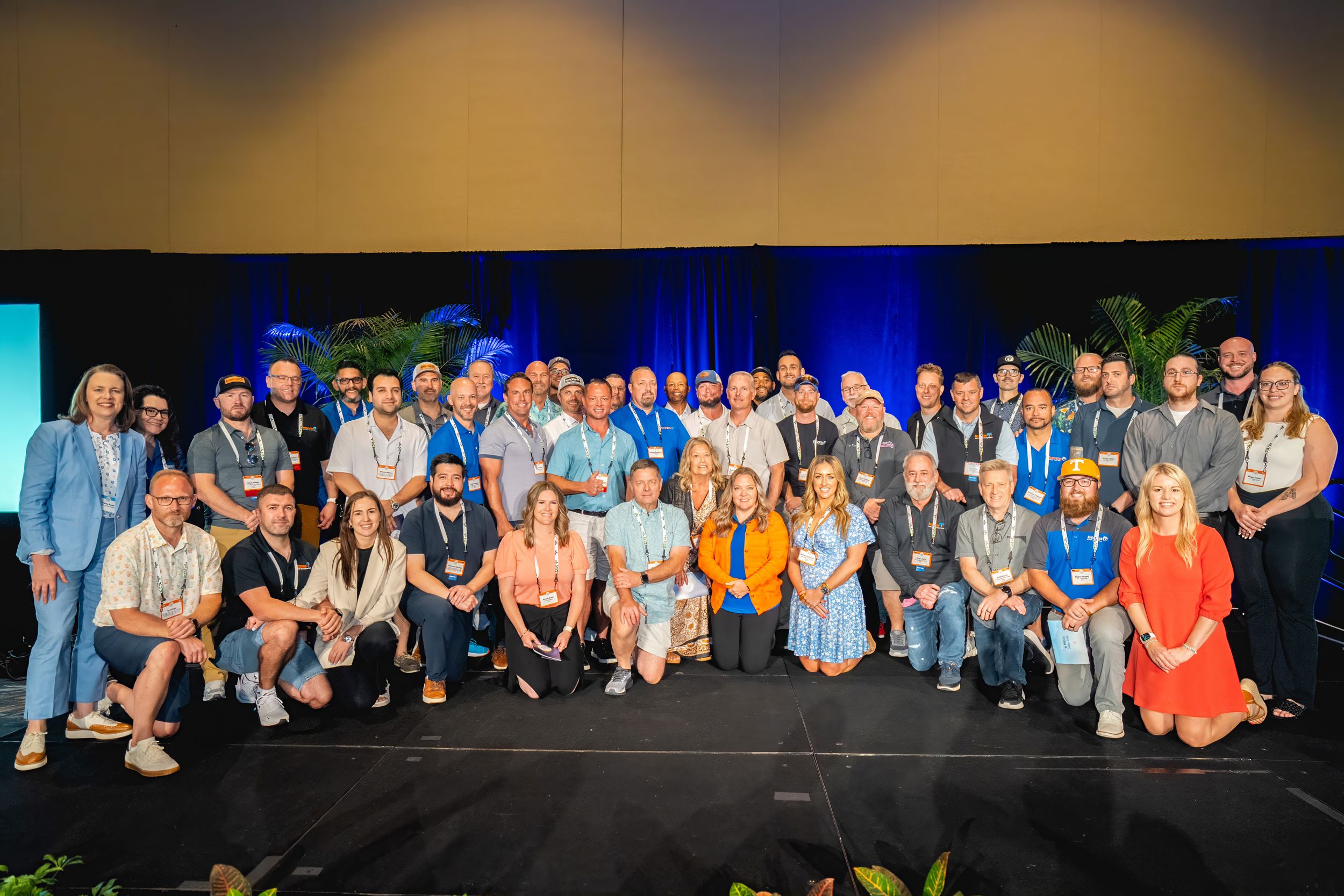 Large group photo of people gathered together on stage with blue lighting and decorative plants for a special event.