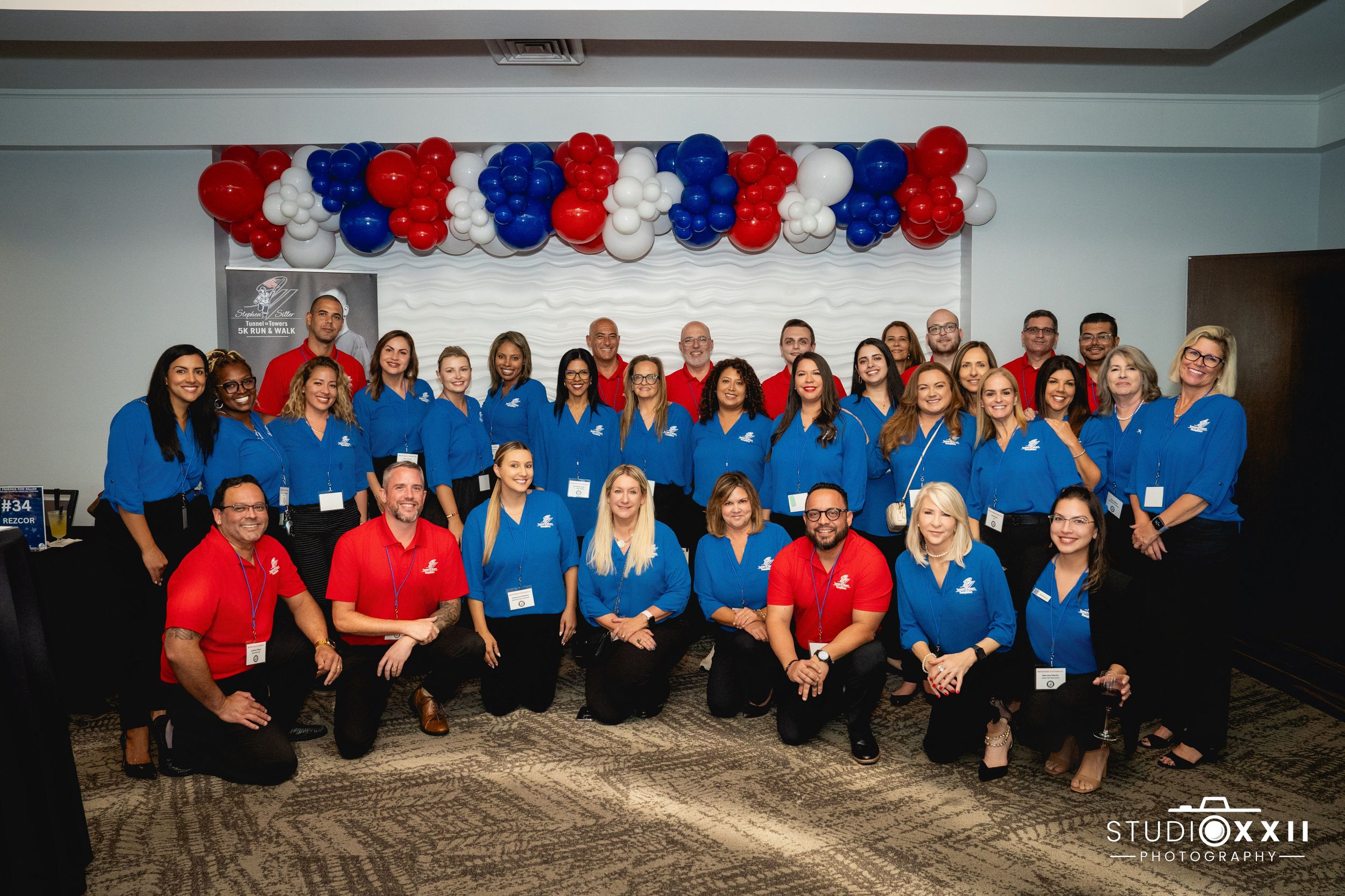 A group of employees in red and blue polo shirts pose together under patriotic balloon decorations at a corporate event.