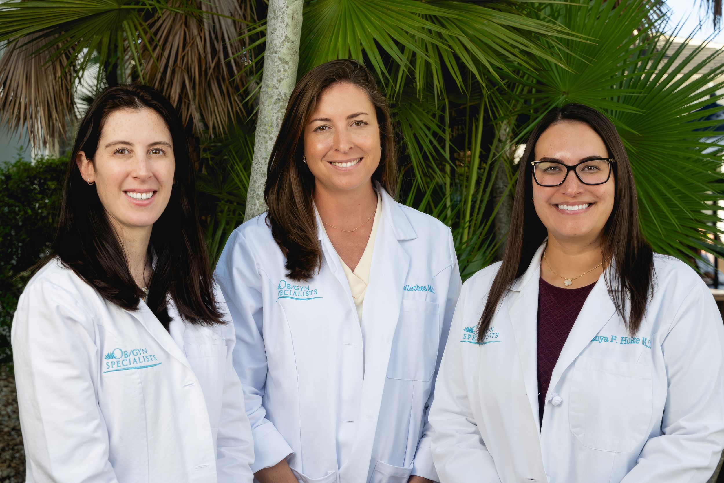 Three healthcare professionals in white lab coats stand together smiling outdoors with palm trees in the background.