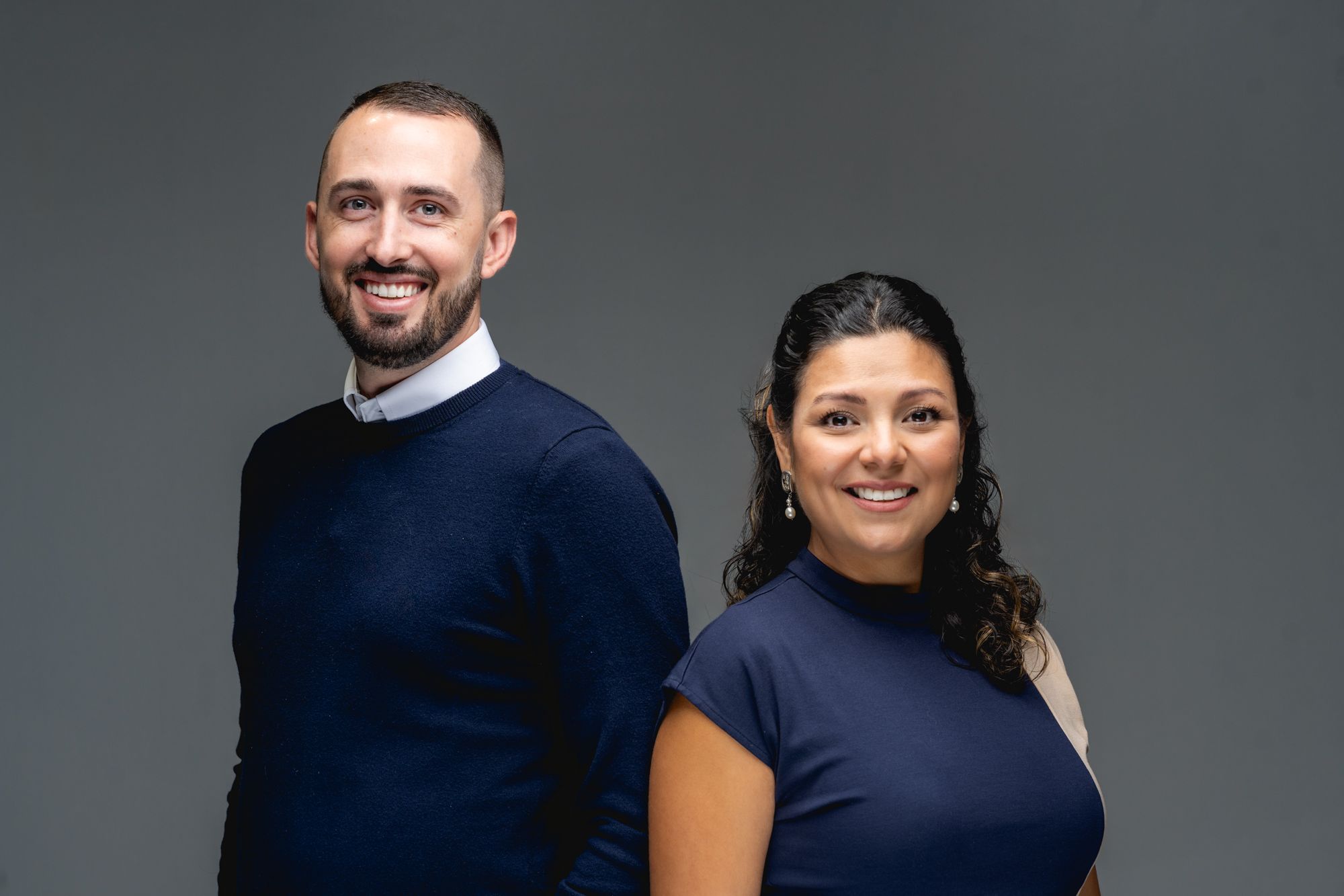 Two business professionals in navy blue tops pose together against a gray studio backdrop.