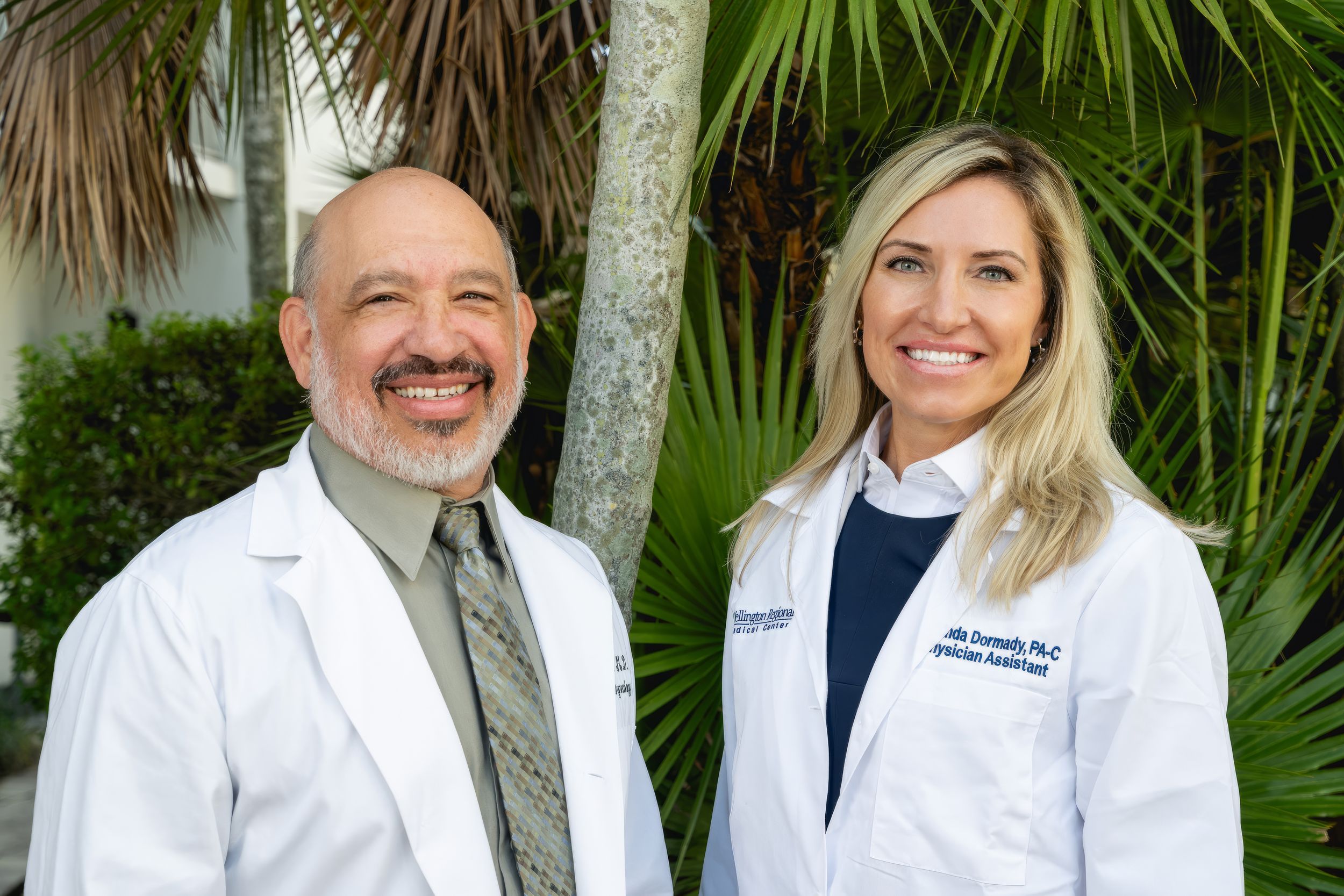 Two medical professionals in white coats stand together smiling in front of tropical palm plants outdoors.
