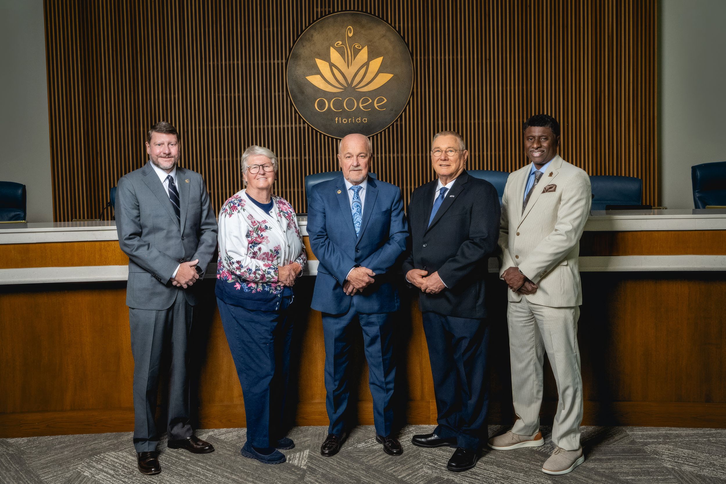 Five professionals pose together in front of a wooden wall with an illuminated logo at a government or corporate venue.