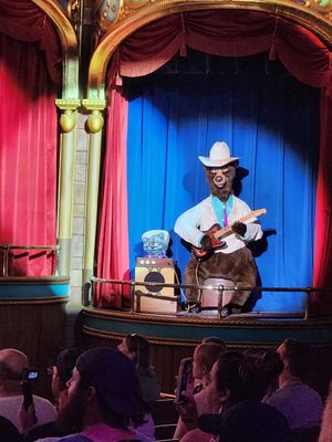 An animatronic performer with a cowboy hat plays music on stage with red curtains and blue backdrop at a theme park attraction.