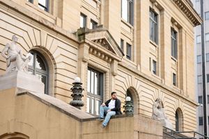 Man sitting on ledge of ornate classical building with stone statues and arched windows.