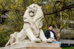 Young man posing beside large stone lion sculpture in urban park setting.