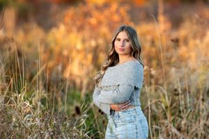 Teen girl in gray sweater and jeans standing in golden autumn field for senior portrait.