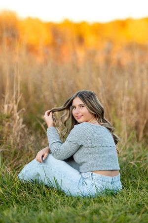 High school senior girl sitting in grass field twirling hair with vibrant fall foliage behind.