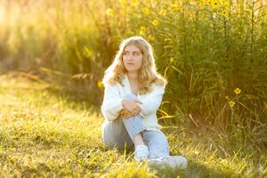 Blonde teenage girl in white sweater and jeans sitting in sunlit golden meadow.