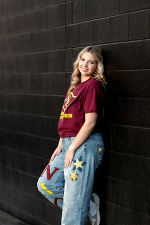 A smiling teenage girl in a maroon school shirt and painted jeans leans against a dark brick wall.