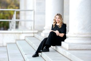 A young blonde woman in an all-black outfit sits gracefully on steps beside tall white columns.