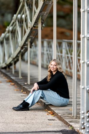 A smiling teenage girl in a black sweater and jeans sits on a metal bridge walkway in autumn.