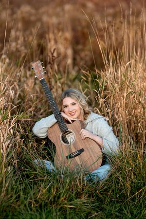 A smiling teenage girl sits in tall golden grass hugging an acoustic guitar in an autumn field.
