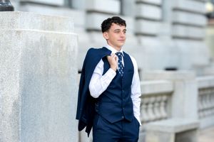 Young man in navy suit carrying jacket over shoulder leaning against stone column.