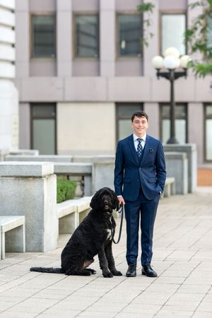 Young man in navy blue suit standing outdoors with black curly-haired dog on leash.