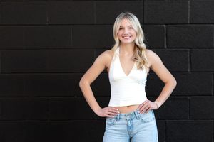 Smiling blonde girl in white crop top and jeans posing against black brick wall.