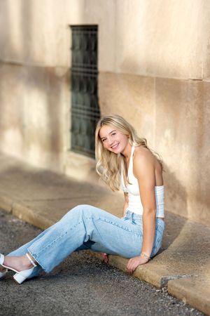 Smiling blonde teen girl in white top and jeans sitting on curb against stone wall for senior photos.