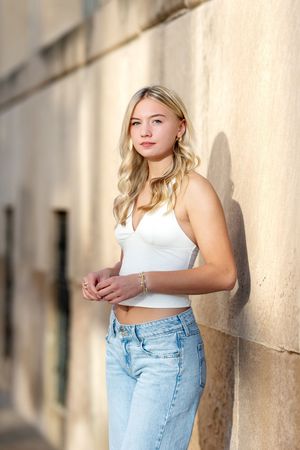 Blonde teenage girl in white crop top and light jeans leaning against stone wall for senior portrait.