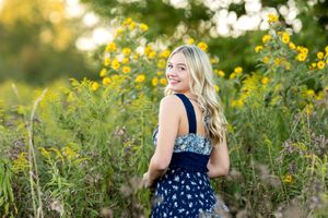Smiling blonde teen girl in navy floral dress standing among yellow wildflowers for senior photos.