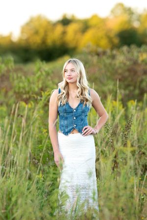 Blonde teen girl in denim vest and white lace skirt standing in tall green meadow for senior portrait.