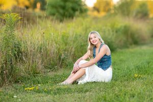 Smiling blonde teen girl in denim top and white skirt sitting on grass in meadow for senior photos.