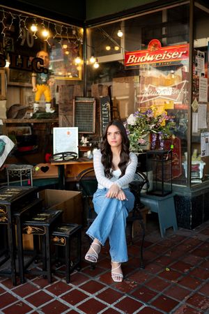 Dark-haired teen girl in white top and jeans sitting outside vintage shop for senior photos.
