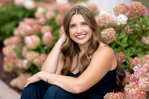 Smiling brunette teen girl in navy outfit sitting among pink hydrangea flowers for senior photos.