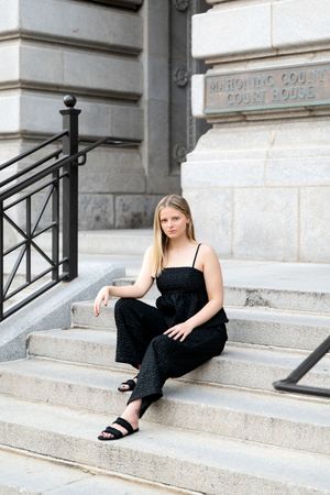 Blonde teen girl in black jumpsuit sits on courthouse steps with stone building in background.