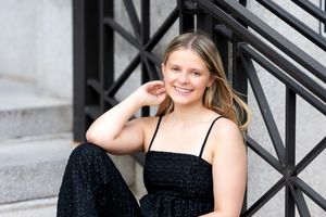 Smiling blonde teenage girl in black outfit sits on steps with iron railing, laughing candidly.