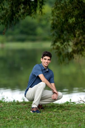 Smiling teenage boy in blue polo shirt crouching by a serene lake under weeping willow branches.