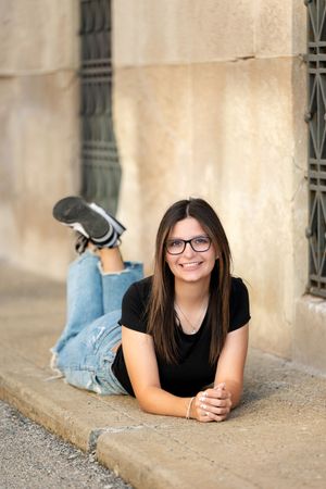 Teenage girl with glasses and dark hair lying on stone steps, smiling in black top and jeans.