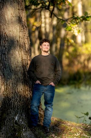 Young man leaning against large tree trunk in sunlit woodland near water.