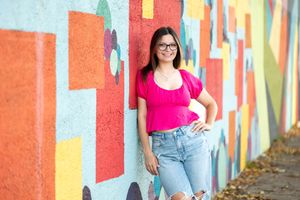 Teenage girl in pink top and ripped jeans smiling against colorful geometric mural wall.