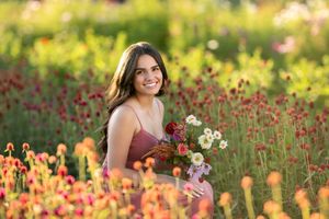 Young woman in pink dress holding wildflowers smiling in a vibrant colorful flower field during golden hour.