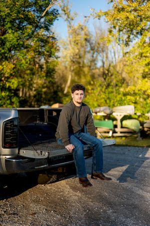 Young man sitting on tailgate of pickup truck in outdoor rural setting.