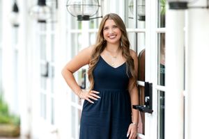 Smiling brunette teen girl in navy dress posing by glass door for senior portrait session.