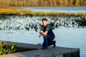 Young man crouching on a wooden dock by a serene lake surrounded by lush greenery during golden hour.