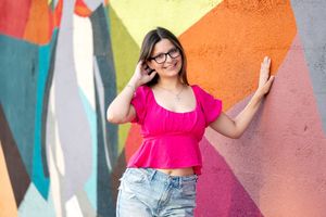 Young woman in pink top and jeans smiling against colorful mural wall wearing glasses.