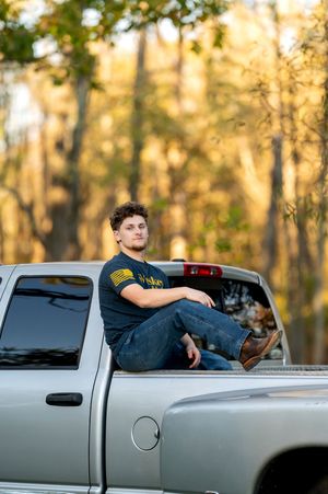 Young man sitting relaxed in the bed of a silver pickup truck with autumn trees in the background.