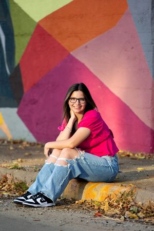 Teen girl with glasses in pink shirt and ripped jeans sitting by vibrant colorful mural.