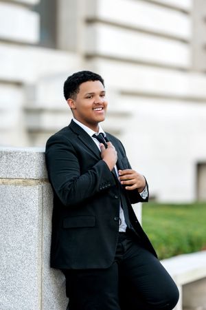 Smiling young man in black suit adjusting tie while leaning against stone pillar outdoors.