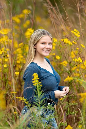 Smiling blonde teen girl in blue cardigan stands among vibrant yellow wildflowers in a field.