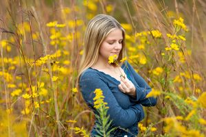 Young blonde girl smelling yellow wildflowers in a golden meadow, wearing a blue long-sleeve top.
