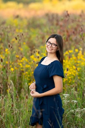 Smiling teen girl with glasses in navy blue dress standing in golden wildflower meadow.