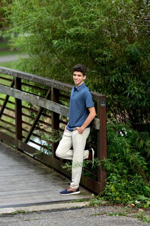Handsome young man in blue polo and khakis leaning on a wooden bridge railing surrounded by greenery.