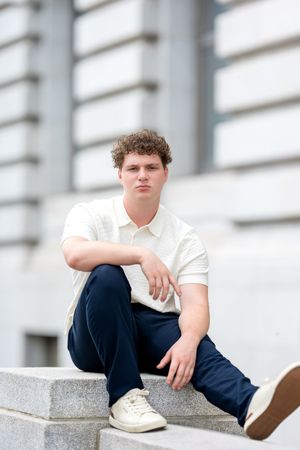 Young man in white polo and navy pants sitting on stone steps outside a classic building.