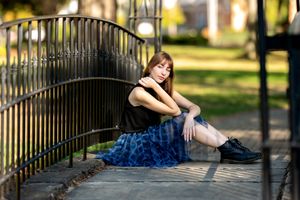 Young woman in a blue skirt and black top sitting against an iron fence in a sunny park.